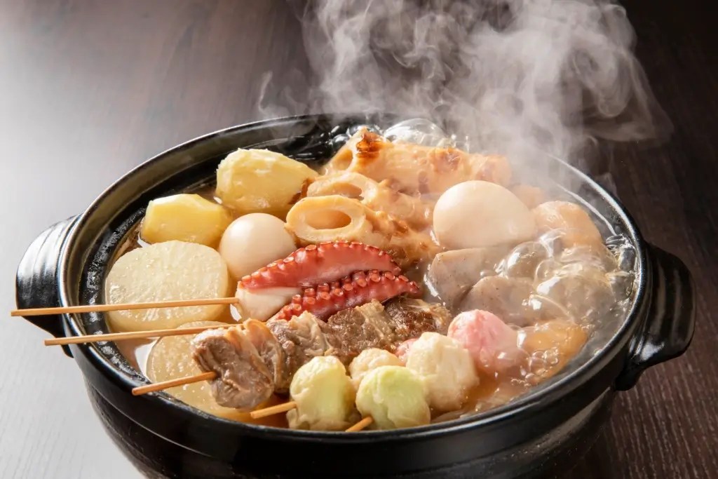 A bowl full of oden stew with dumplings, skewers and other foods.