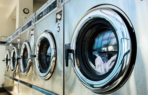 Several stainless steel front-loading washing machines lined up in a laundromat or commercial setting. One machine in the foreground reveals clothes inside.