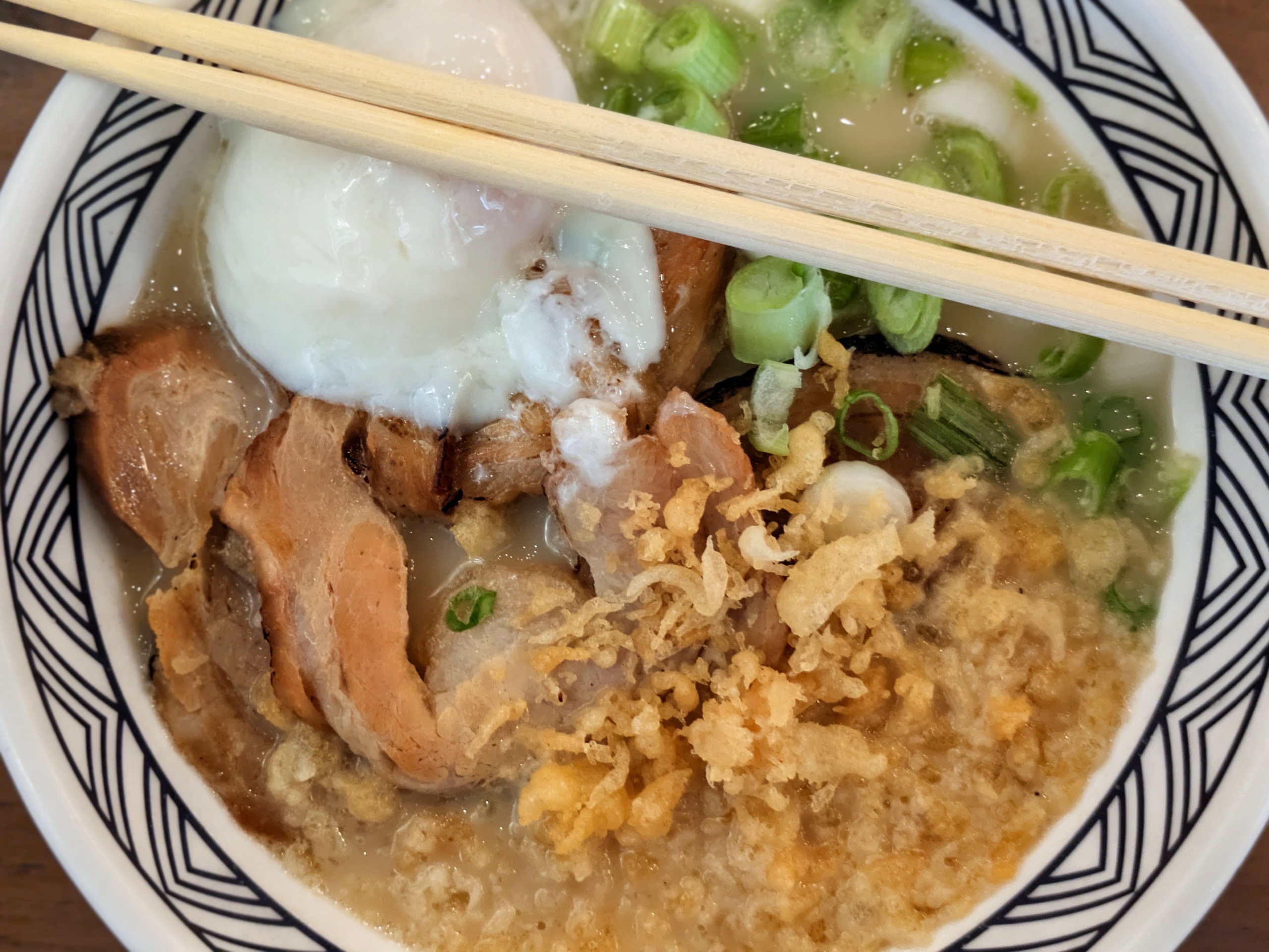 A pair of wooden chopsticks rests across a patterned bowl filled with udon, topped with a soft egg, sliced pork, green onions, and crispy fried bits in a creamy broth.