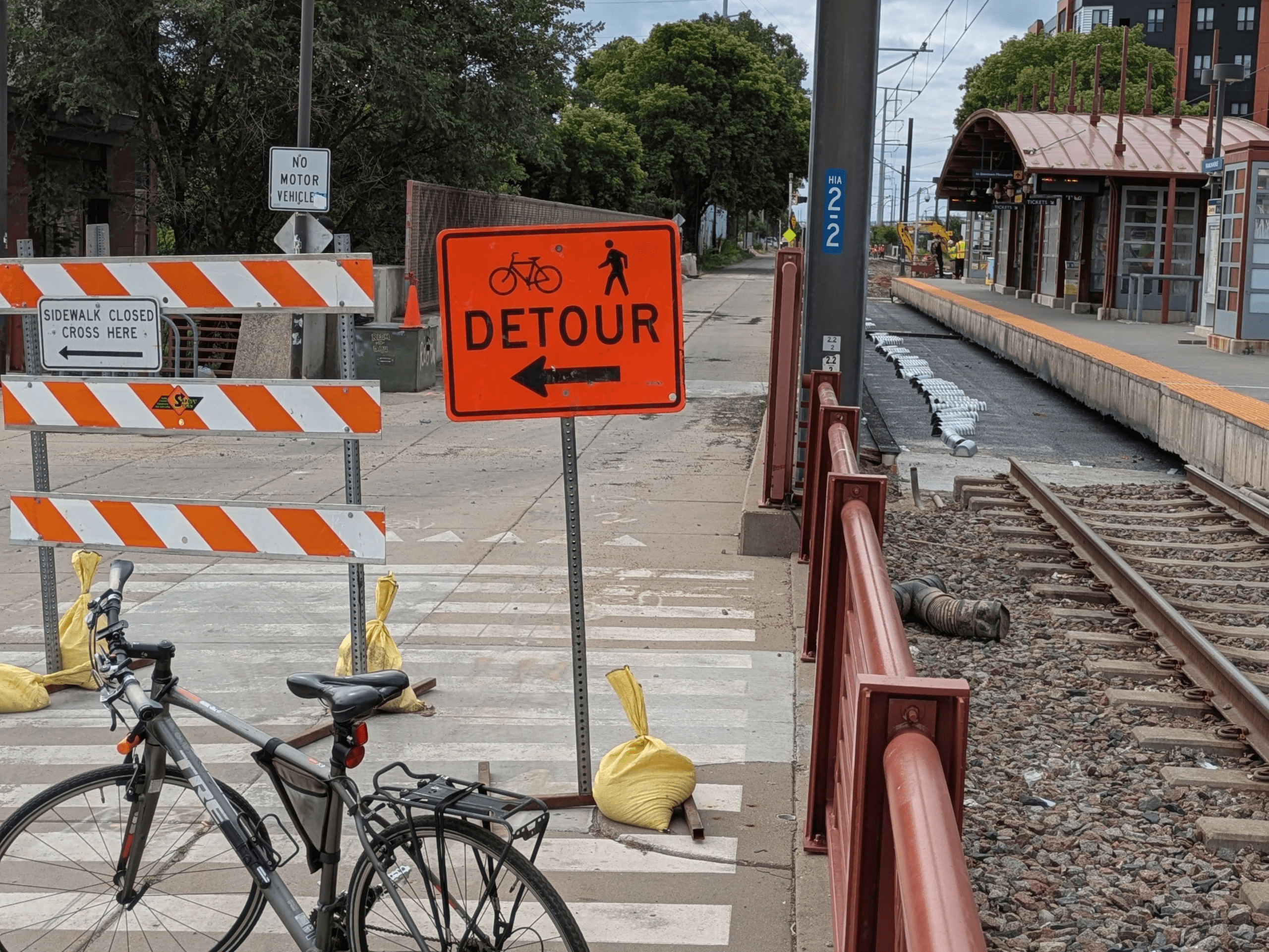 A bicycle is parked in the foreground near orange and white construction barriers and a detour sign, with railroad tracks with a section of track missing, and a train station platform visible in the background.