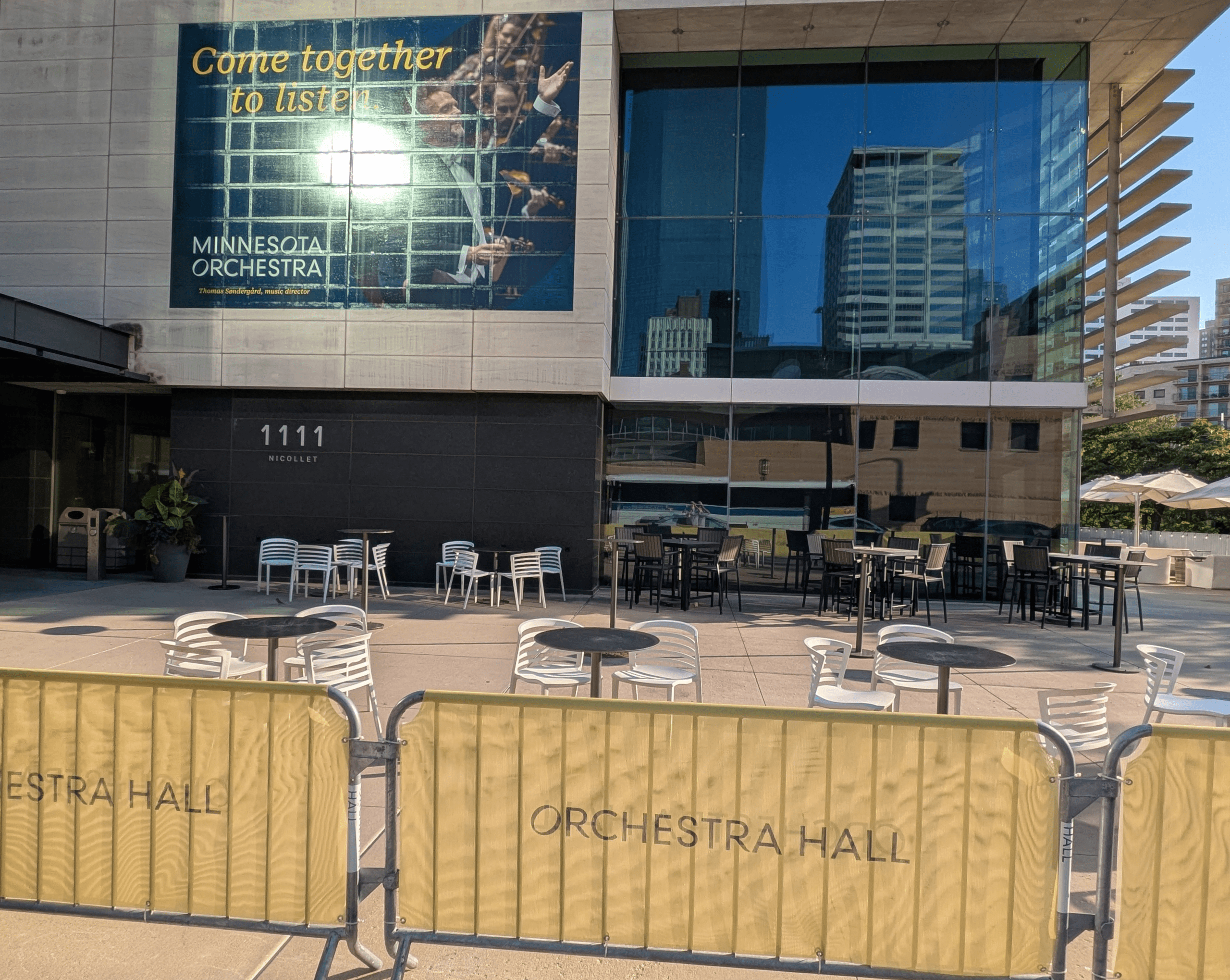 A modern building featuring a Minnesota Orchestra poster and reflective blue glass windows stands above an outdoor patio area filled with numerous tables, chairs, and umbrellas, with yellow mesh barriers displaying Orchestra Hall in the foreground.
