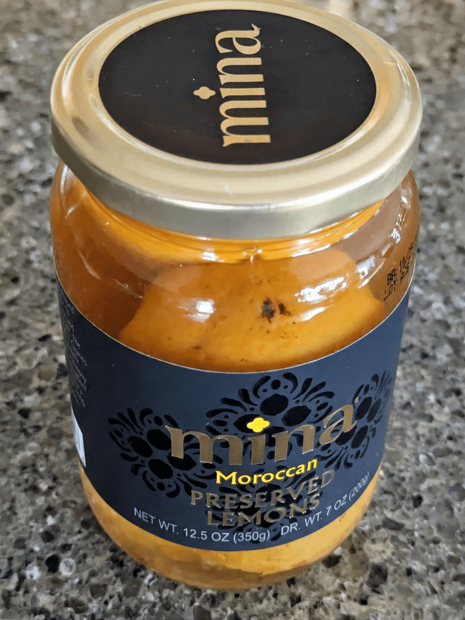 A jar of Mina Moroccan preserved lemons with a gold lid and black label sits on a speckled gray countertop.