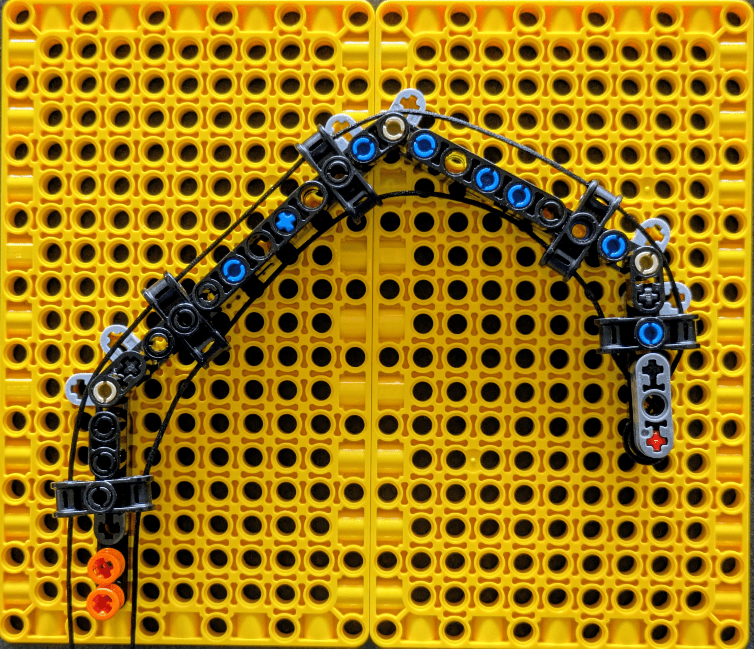 A curved assembly of black, blue, white, gray, and orange LEGO connected by a black string rests on a bright yellow grid board with many circular holes.
