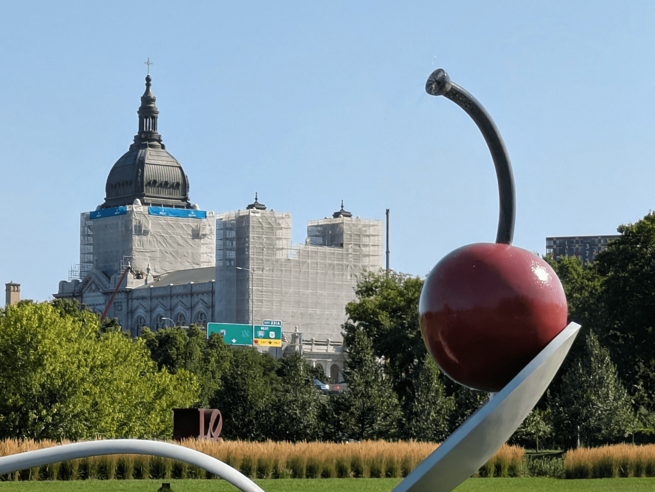In a park under a clear blue sky, a large red cherry sculpture sprays water from its stem while a grand building covered in scaffolding rises in the background among trees.