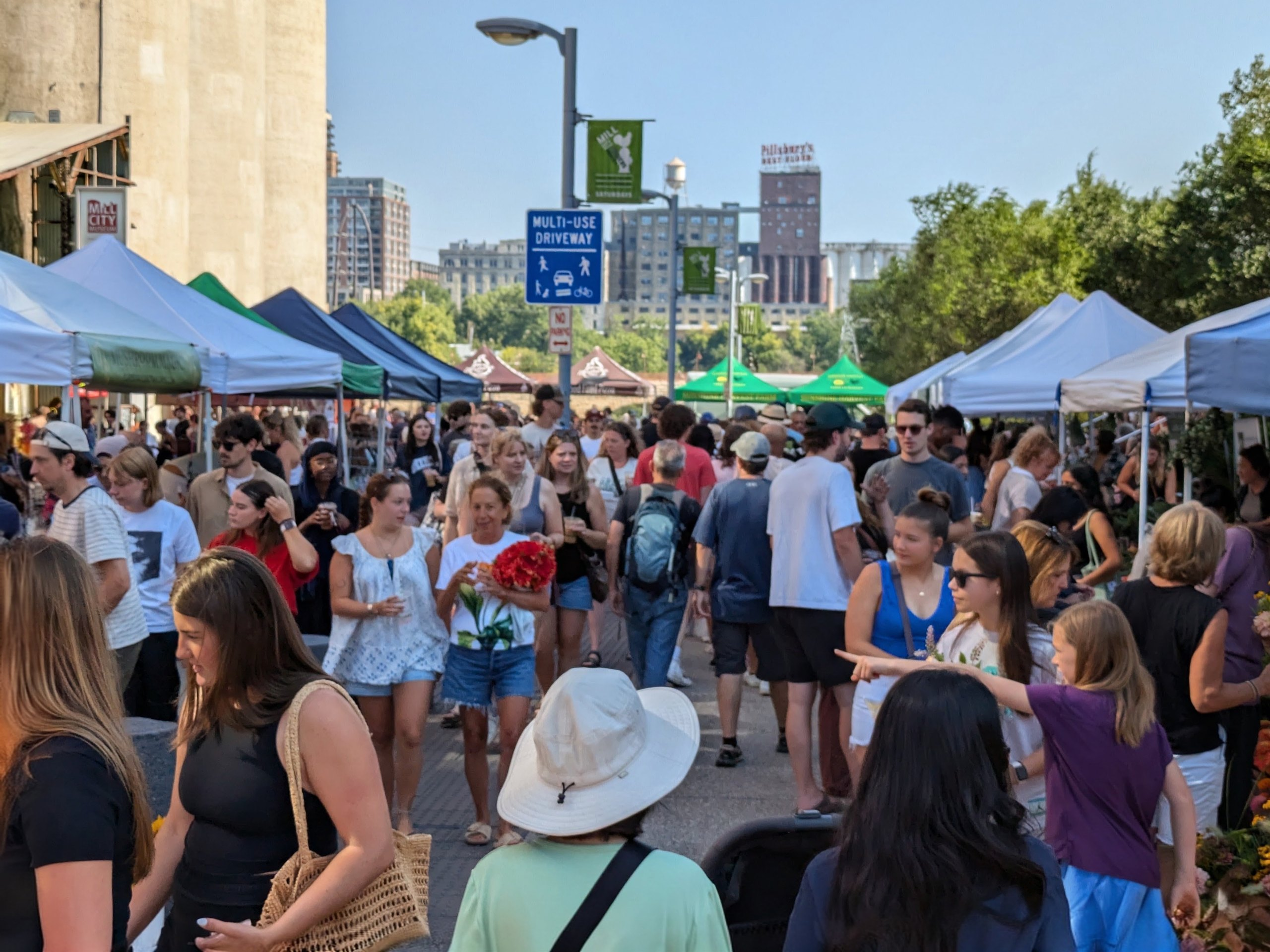 Crowds of people stroll through an outdoor market lined with tents, set against a backdrop of city buildings, including a prominent Pillsburys sign, under a clear blue sky.