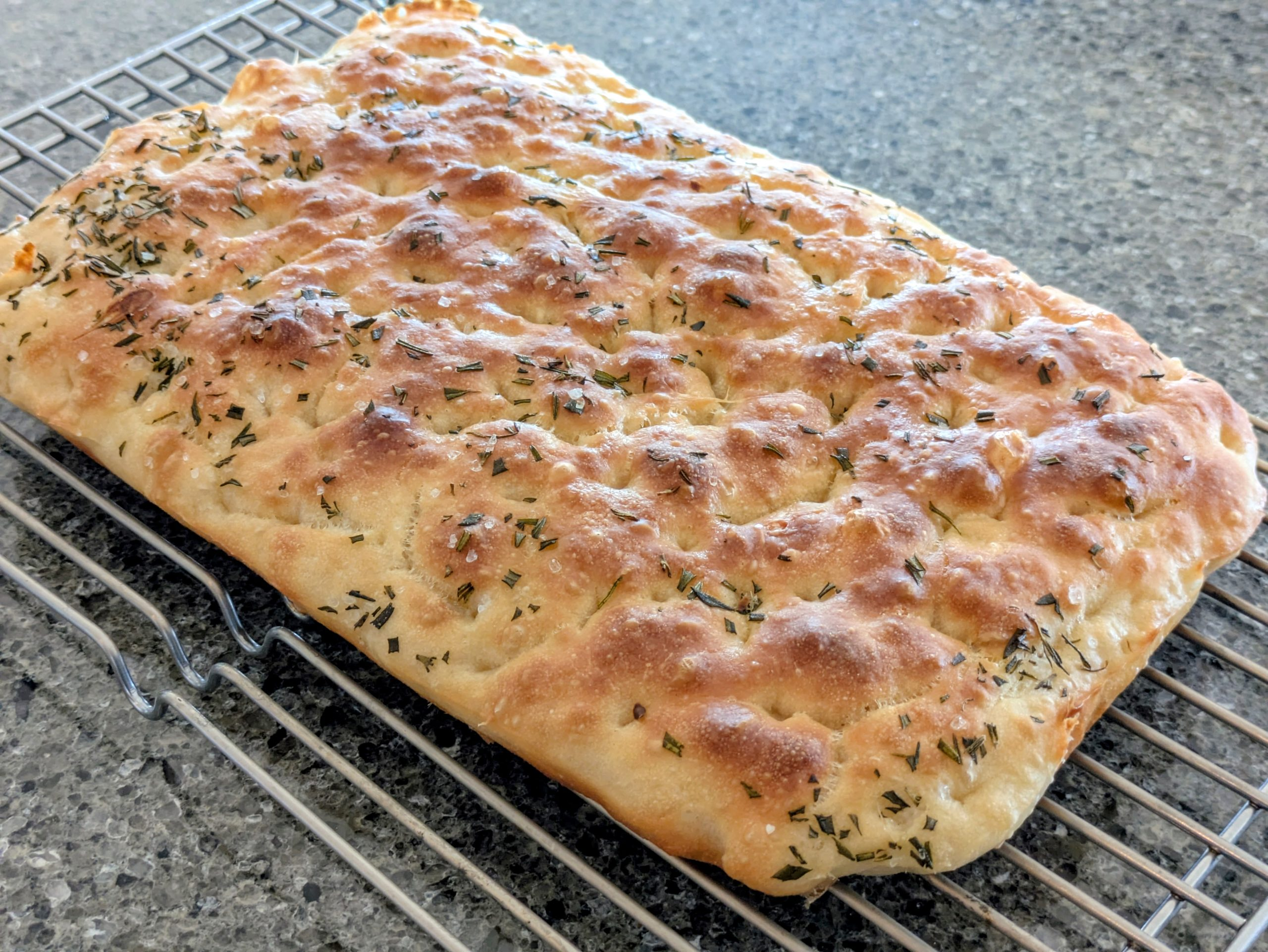 A golden brown focaccia bread topped with herbs and salt rests on a wire cooling rack over a speckled gray countertop.