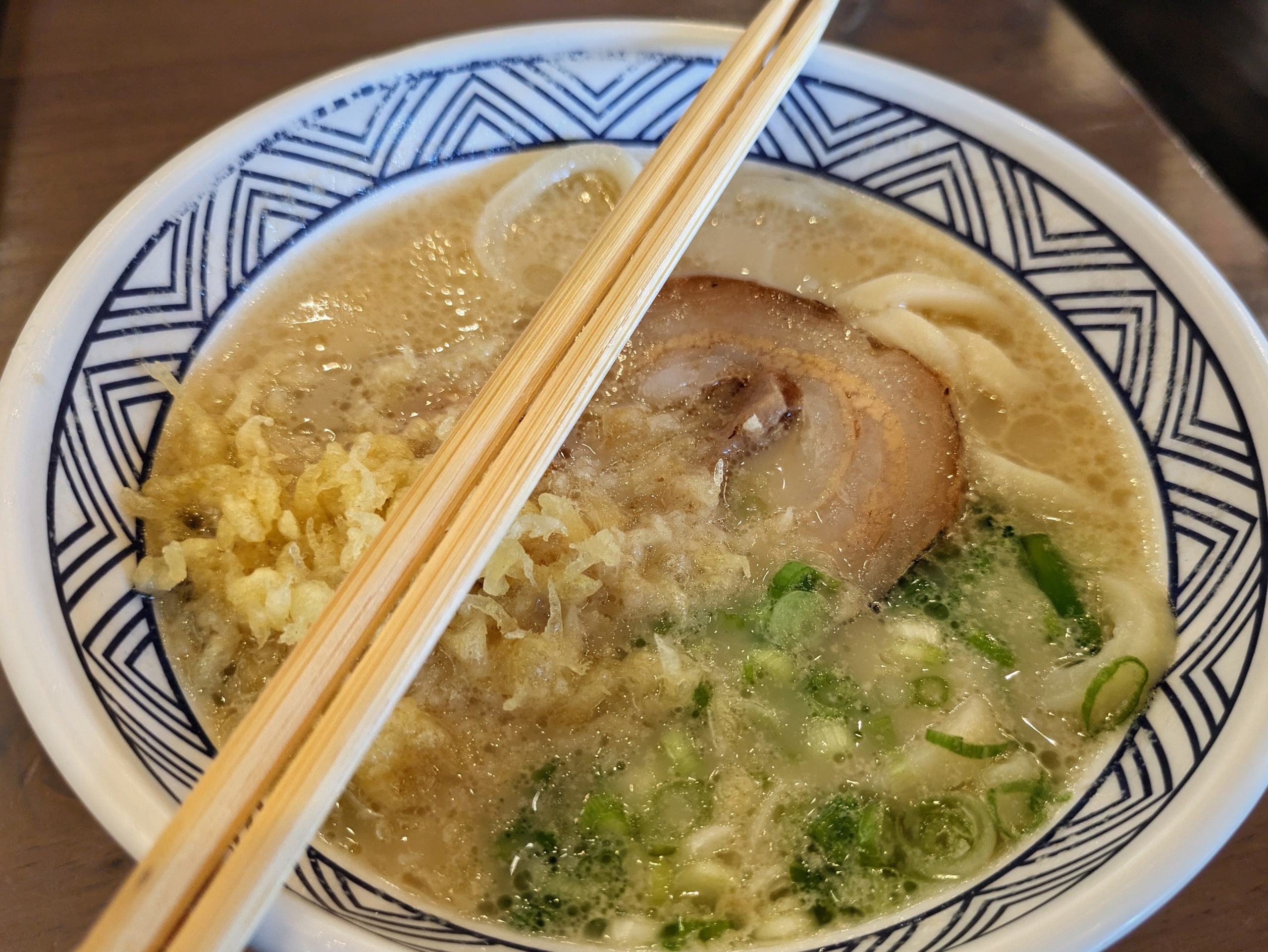 Wooden chopsticks rest on a white bowl filled with udon noodles, a slice of pork, green onions, and tempura flakes in a savory broth.