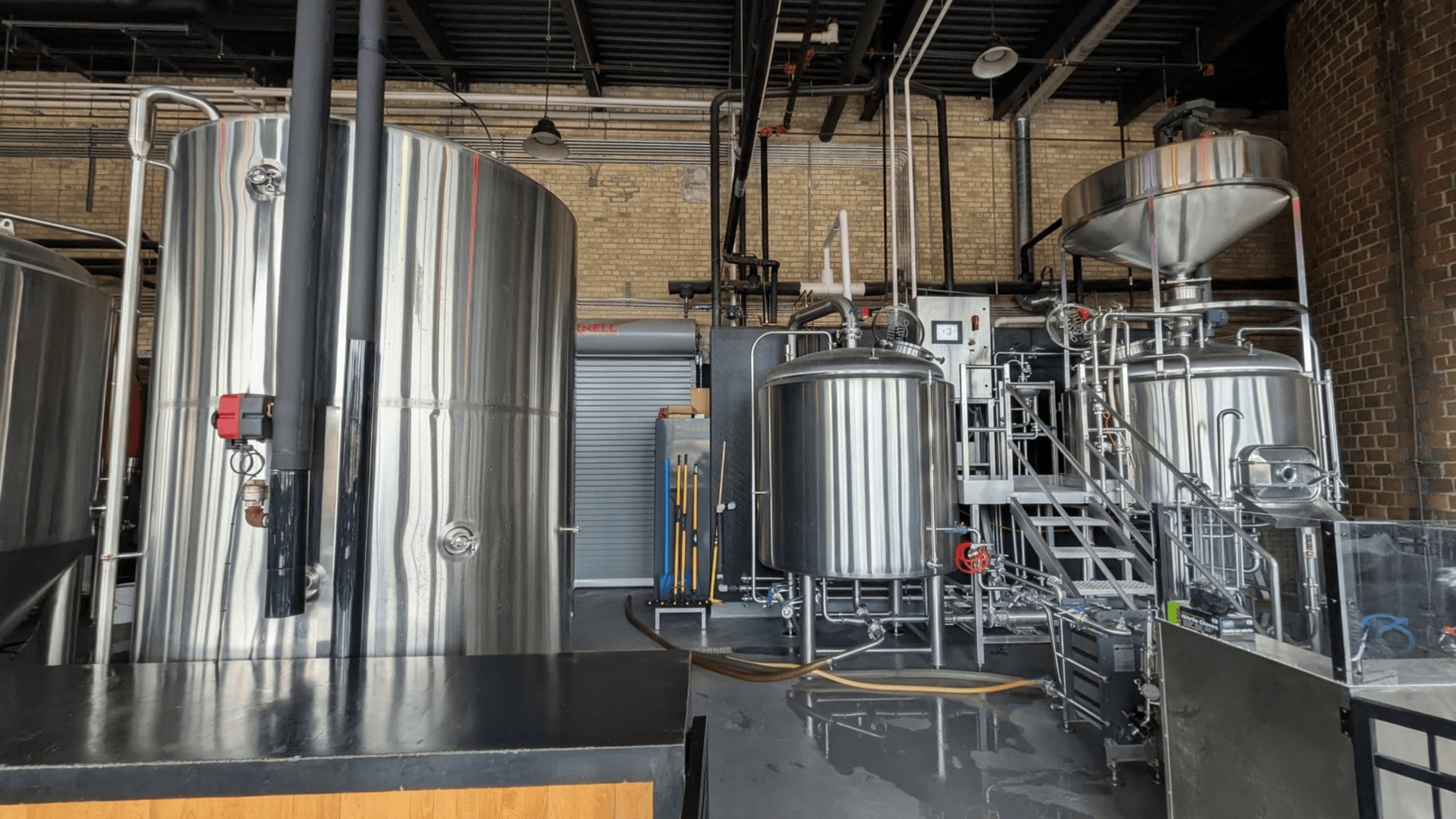 A modern industrial interior showcases multiple large, gleaming stainless steel tanks, an elaborate system of pipes, and control panels, all set against a backdrop of brick walls and a dark ceiling structure.