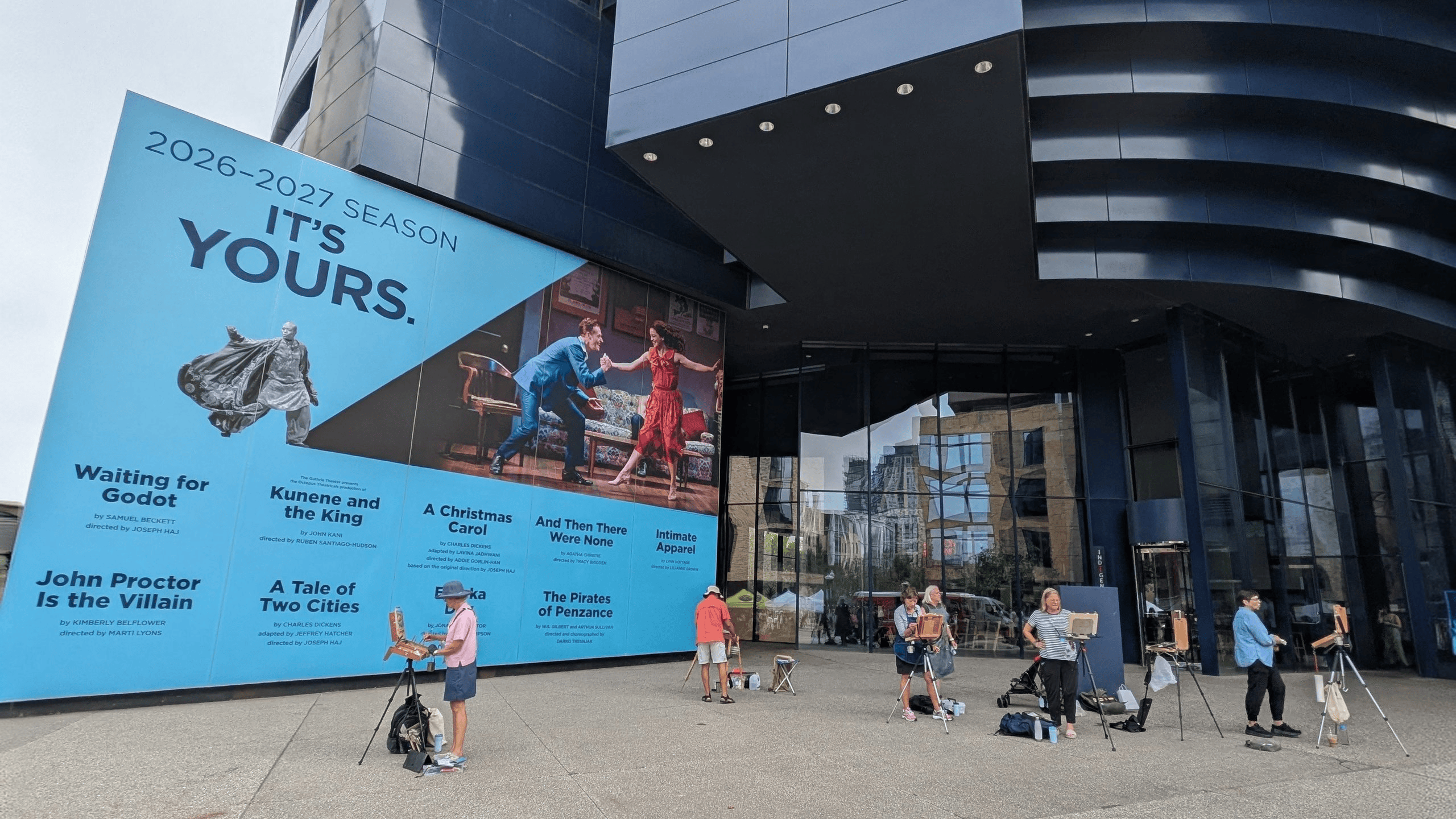 A prominent blue billboard displaying the 2026 2027 theater season schedule and play images is attached to a modern dark building, while several people paint on easels on the ground below.