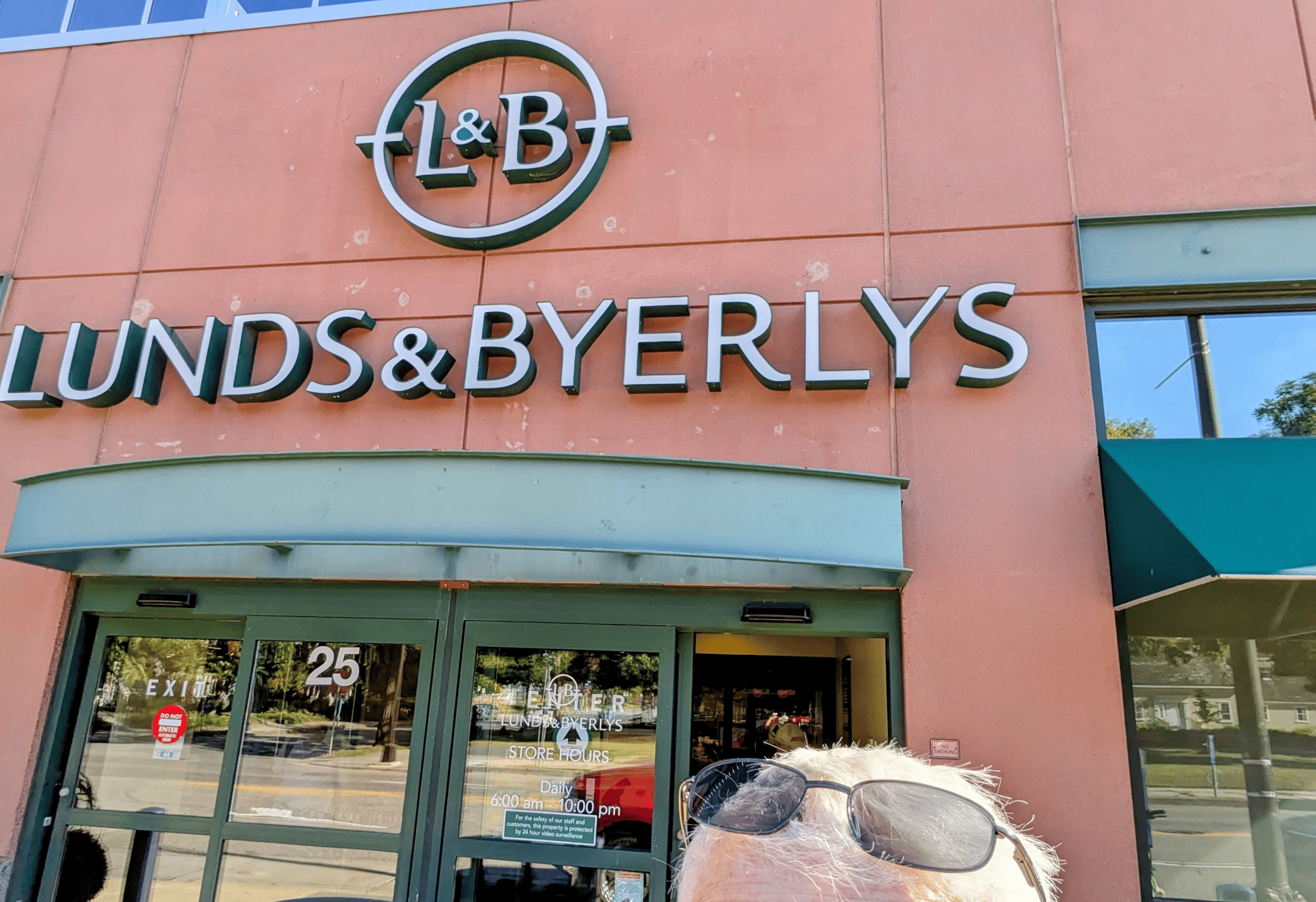 The salmon colored facade of a Lunds and Byerlys grocery store is shown with its green framed glass entrance doors reflecting a street scene, while the top of a persons head wearing sunglasses is visible at the bottom right.