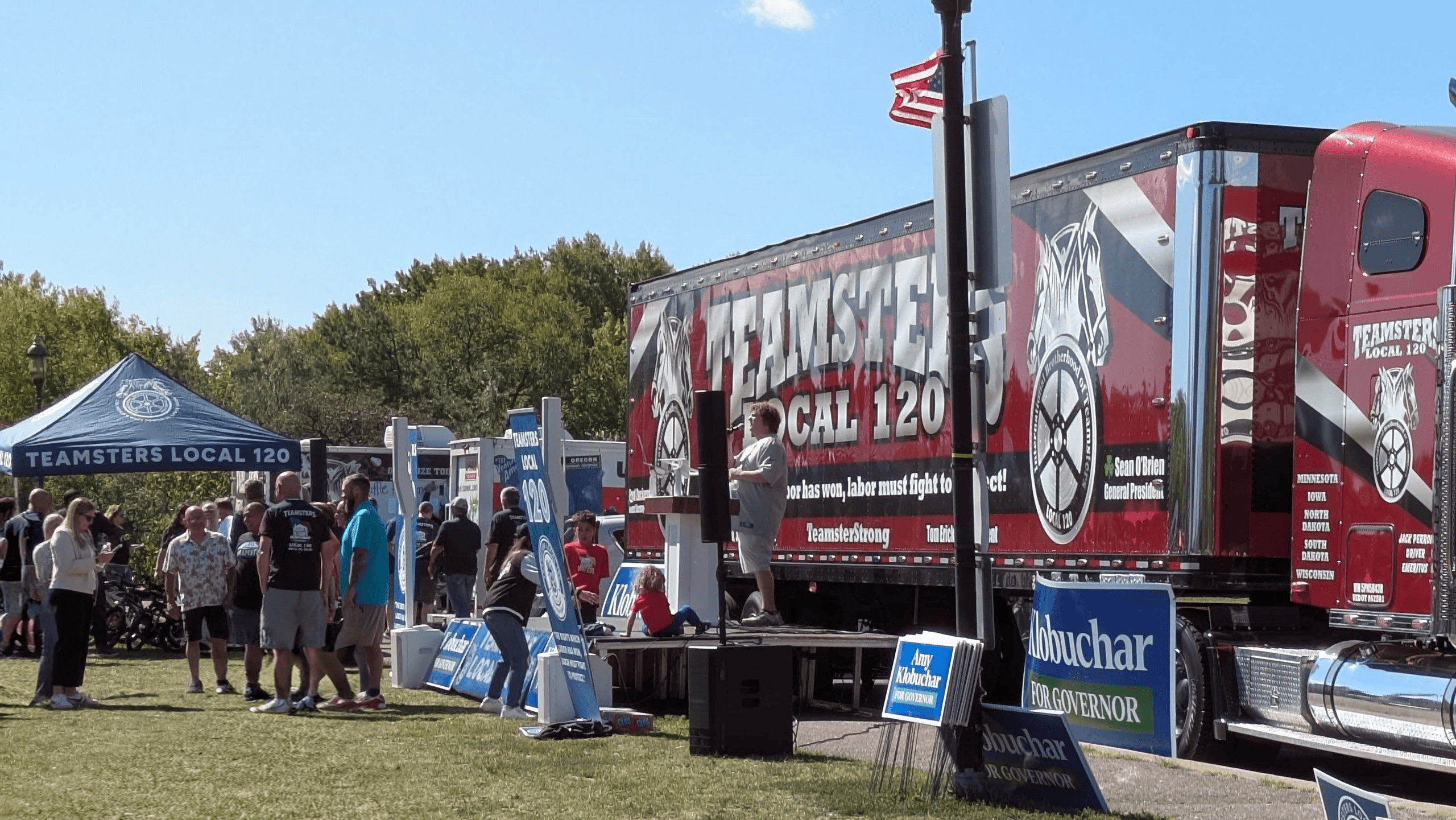 A crowd of people gathers outdoors on a sunny day next to a large Teamsters Local 120 truck and canopy tent, with political campaign signs in the foreground.