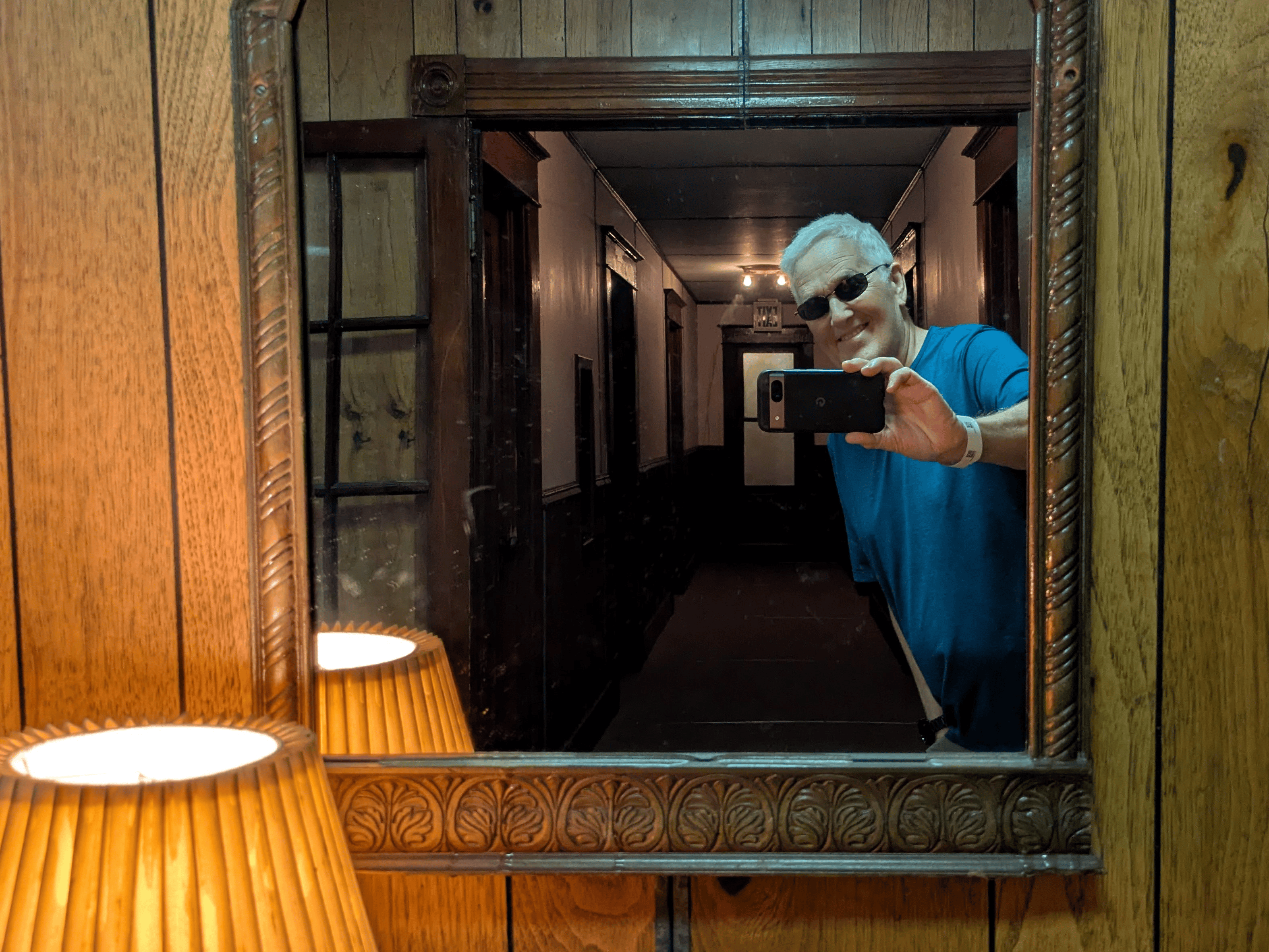A smiling man wearing sunglasses and a blue shirt takes a mirror selfie with his black phone, the ornate framed mirror reflecting a dimly lit hallway with a ceiling fixture and a distant door, as a pleated lampshade is visible in the bottom left against a wood paneled wall.