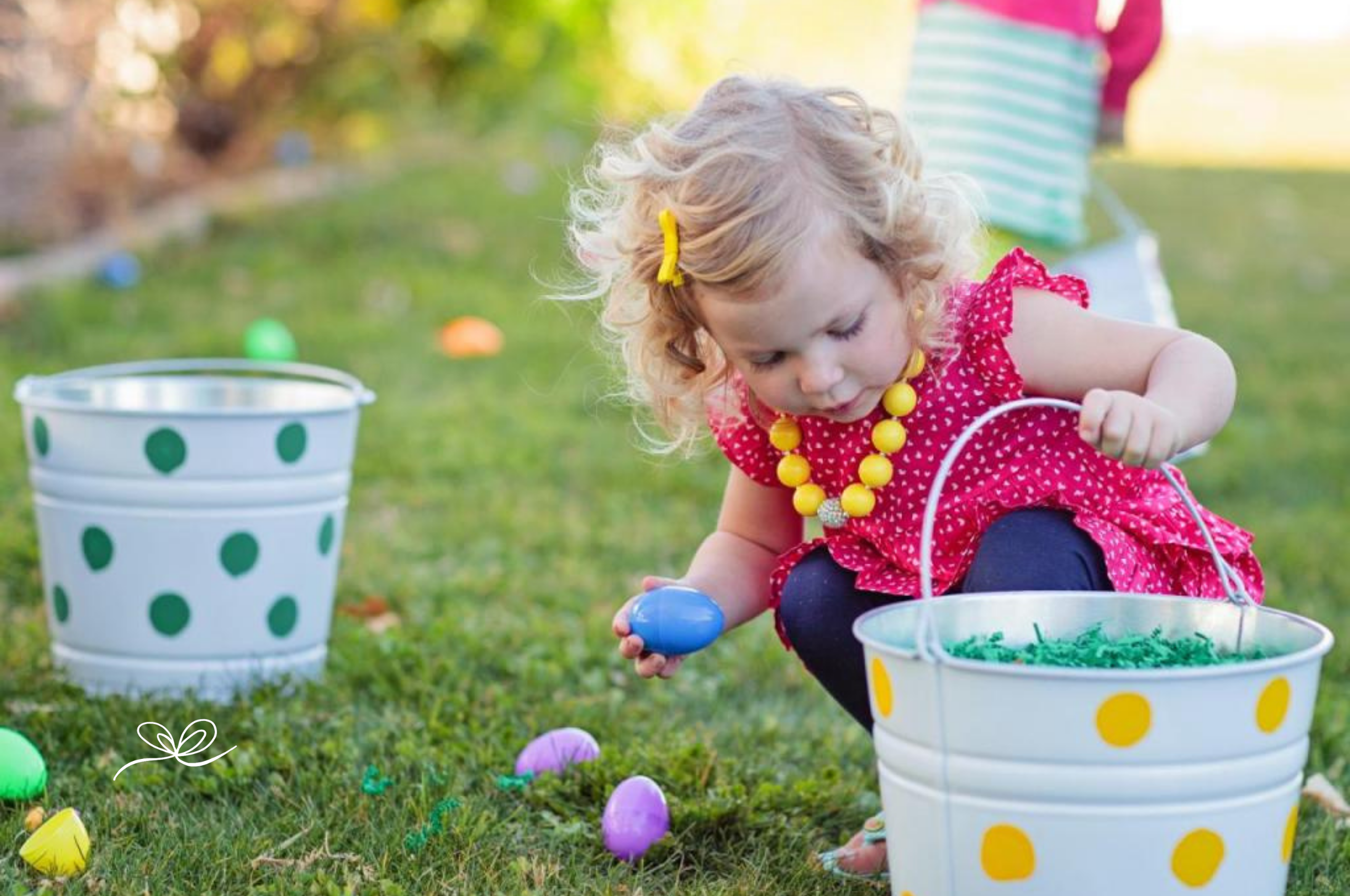 Toddler in a pink dress collecting plastic Easter eggs on a lawn at an Easter egg hunt