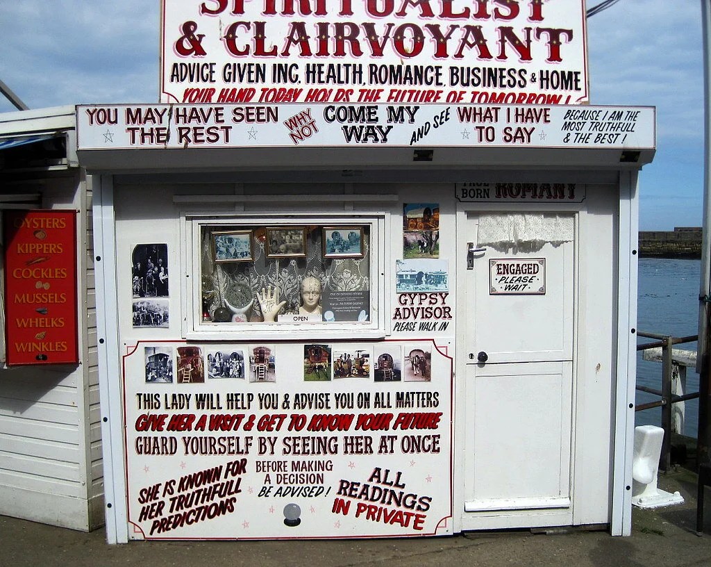 A seaside fortune teller's booth covered in hand-painted signs: "Spiritualist & Clairvoyant," "Get to know your future" and "All readings in private"