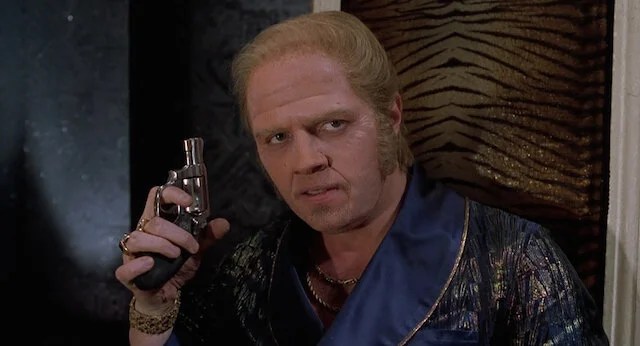 Scene from Back to the Future Part II of a man in a silk robe and gold jewelry holding a revolver