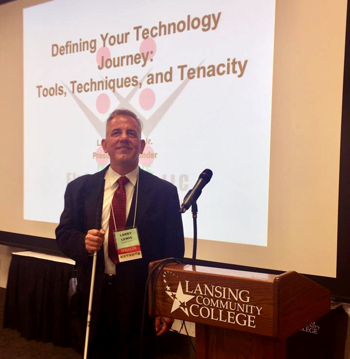 Flying Blind, LLC's Founder, Larry Lewis, in suit and tie prior to conducting a presentation, stands smiling in front of a large, wall-sized projection of the Flying Blind, LLC Logo.