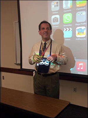 Photo of Flying Blind, LLC's President, Larry Lewis, conducting a presentation in front of a large screen upon which is being projected the screen of an iOS device which he is controlling using a small refreshable Braille display that is hanging, otherwise untethered, around his neck.