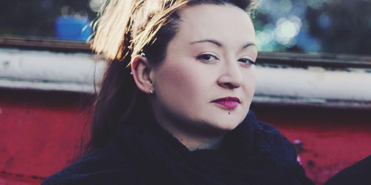 A portrait of folk singer Eliza Carthy, seated outside with a thoughtful expression, wearing a black scarf and a lip piercing, with sunlight highlighting her hair.