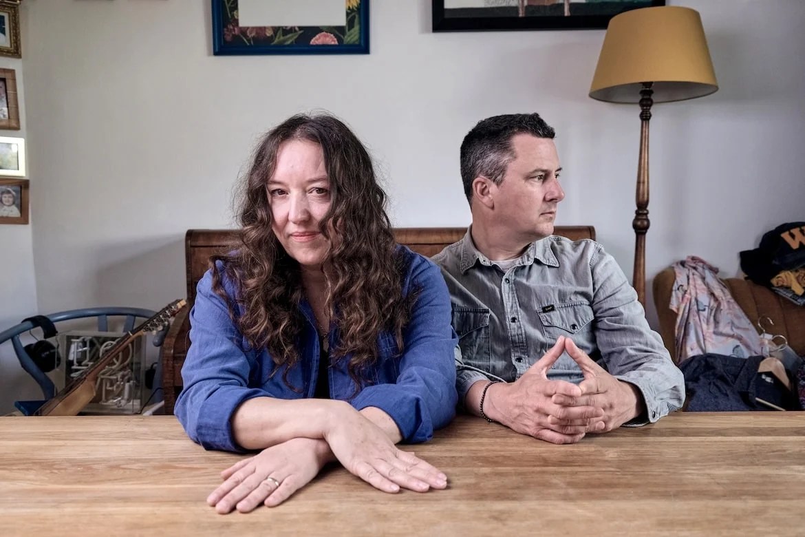 An image featuring Kathryn Williams and Withered Hand sitting at a wooden table. Williams, on the left, is resting her arms on the table and looking directly at the camera with a faint smile, her long, wavy hair framing her face. Withered Hand, on the right, is seated beside her, hands clasped together and gazing thoughtfully to the side. Behind them is a cozy room with a lamp and framed pictures on the wall.