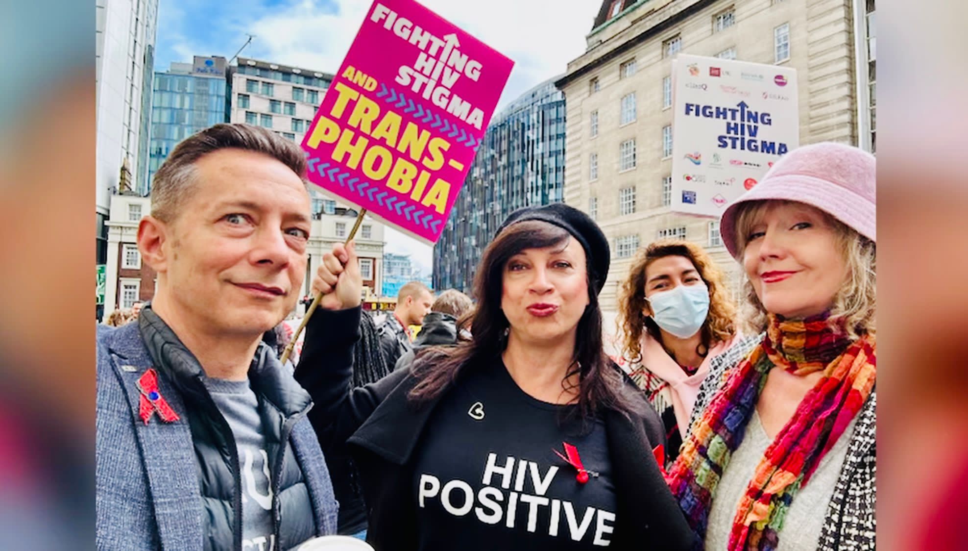 Photo of Victoria, a Latina British trans woman with long brown hair. She's wearing a t-shirt which says HIV positive and is at a demo with some other women and a man.
