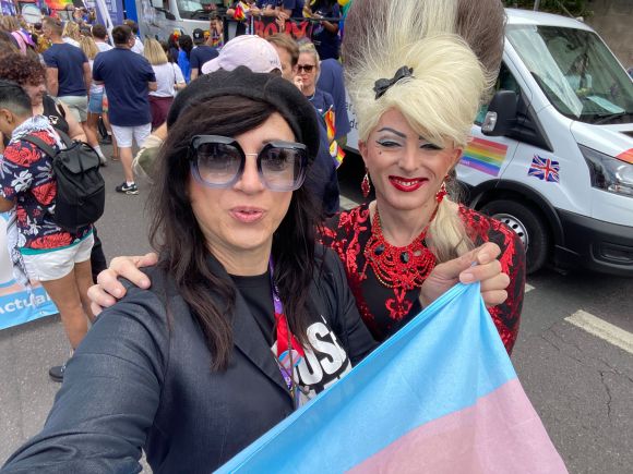 Photo of Victoria, a Latina British trans woman with long brown hair. She's holding a trans flag and posing for a photo with a drag queen.