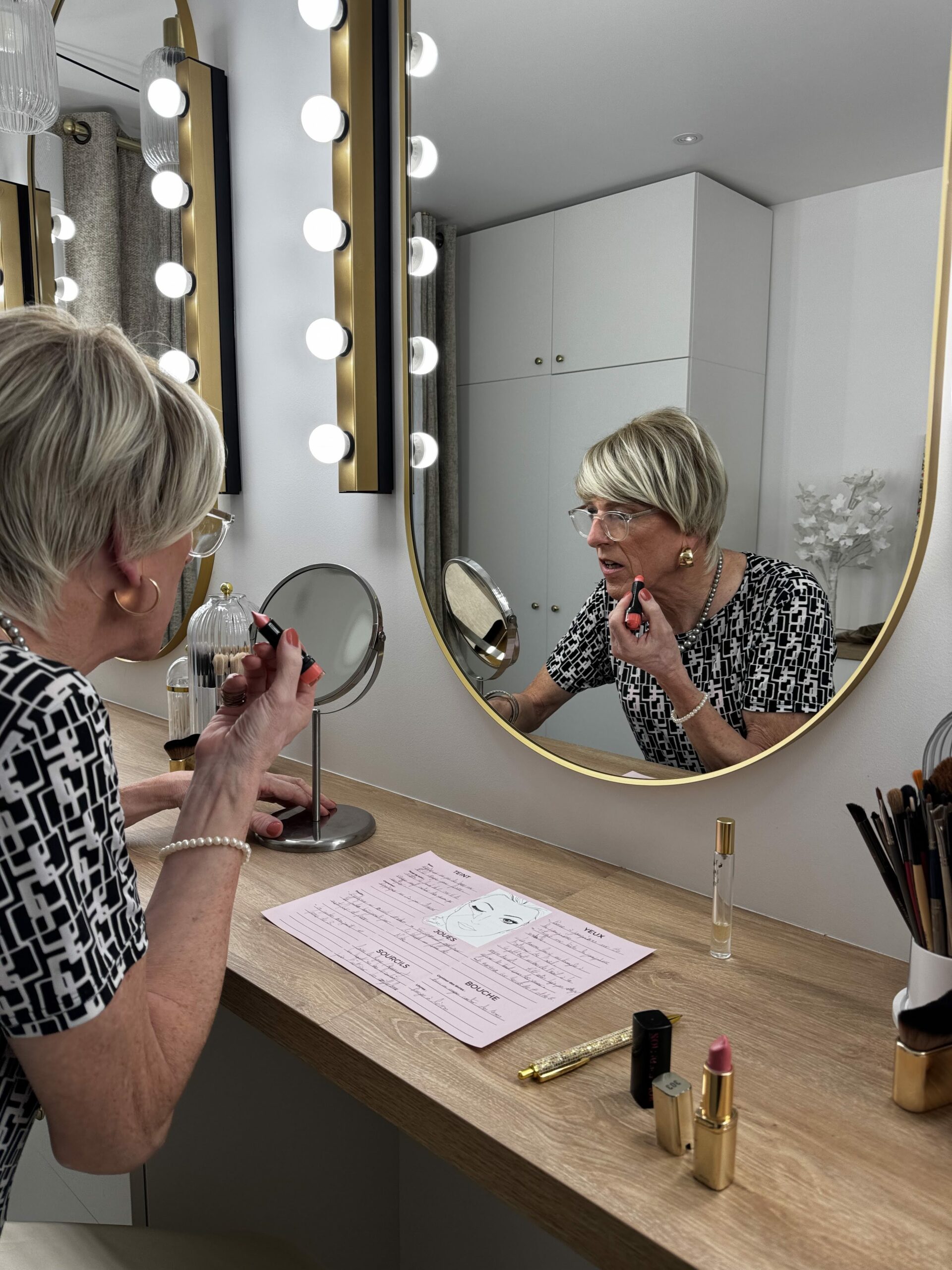 Une personne transgenre ou travestie applique soigneusement du maquillage devant un miroir illuminé. L'espace est aménagé avec des produits de beauté et des outils de maquillage, reflétant une routine soignée et affirmée