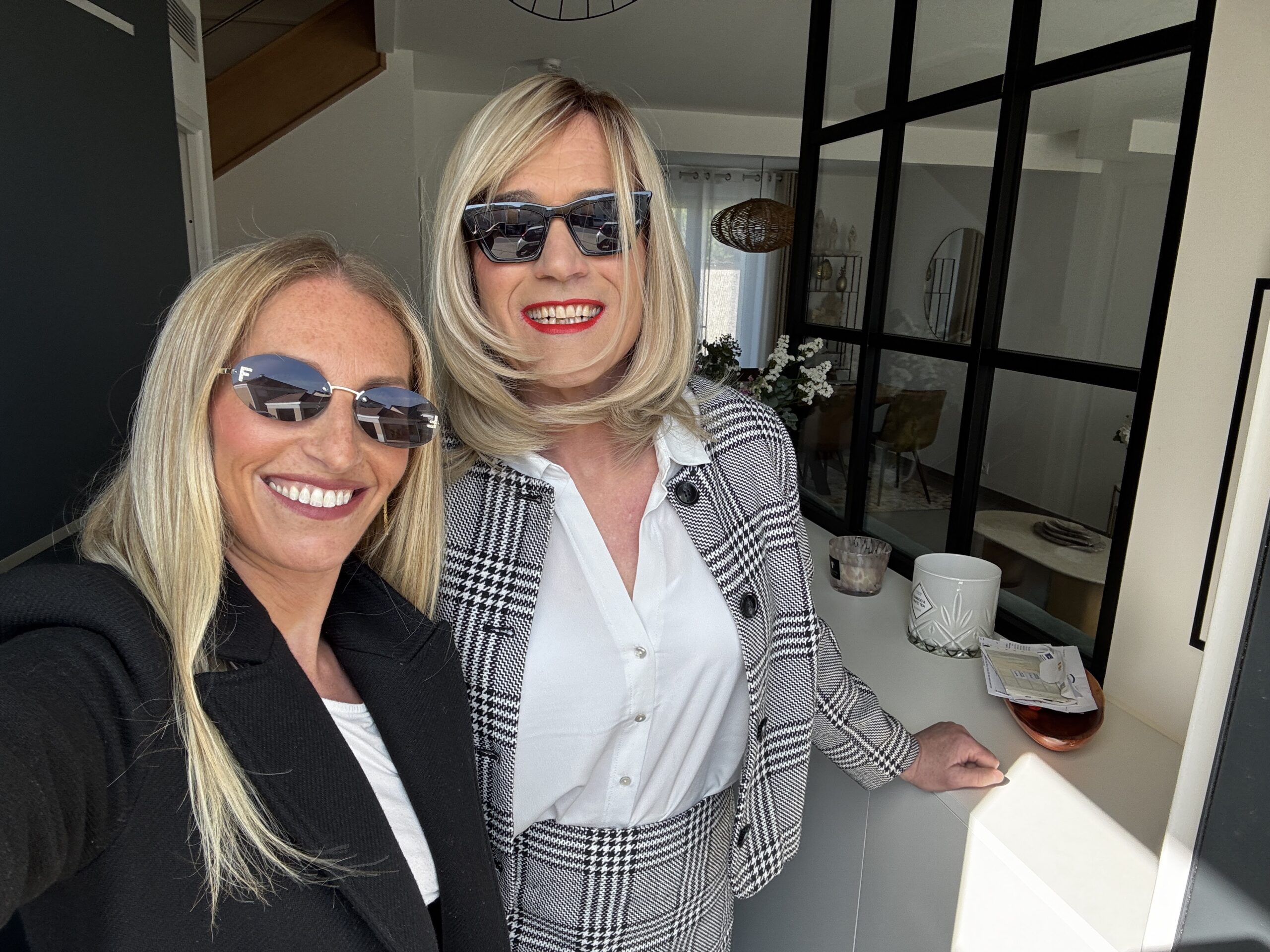 Jennifer et Lucille chez Transbeauté prennent un selfie dans un intérieur moderne. Jennifer porte un tailleur pied-de-poule noir et blanc, une perruque blonde et des lunettes noires. Lucille porte un blazer noir et des lunettes de soleil. Toutes deux sourient avec éclat.