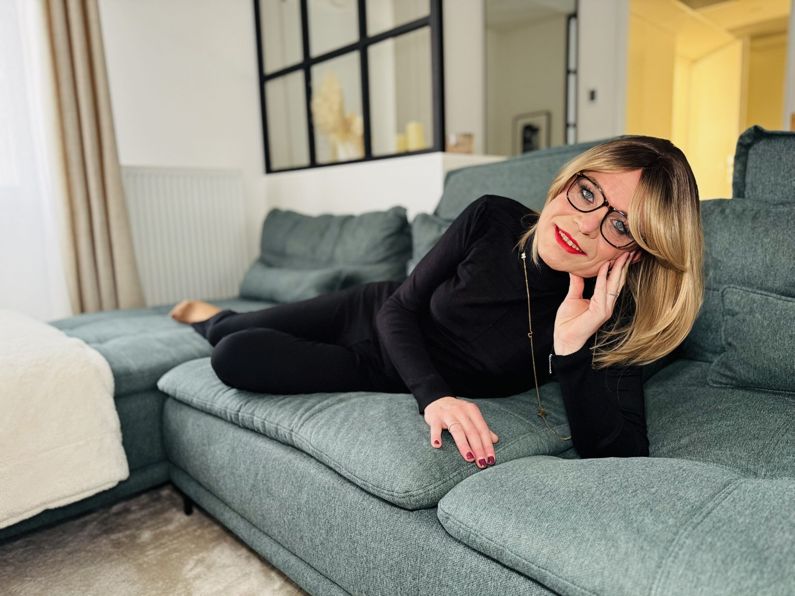 Femme transgenre allongée sur un canapé vert d’eau, vêtue de noir, avec des lunettes et des cheveux blonds lissés. Elle sourit doucement, dans un intérieur lumineux et apaisant.