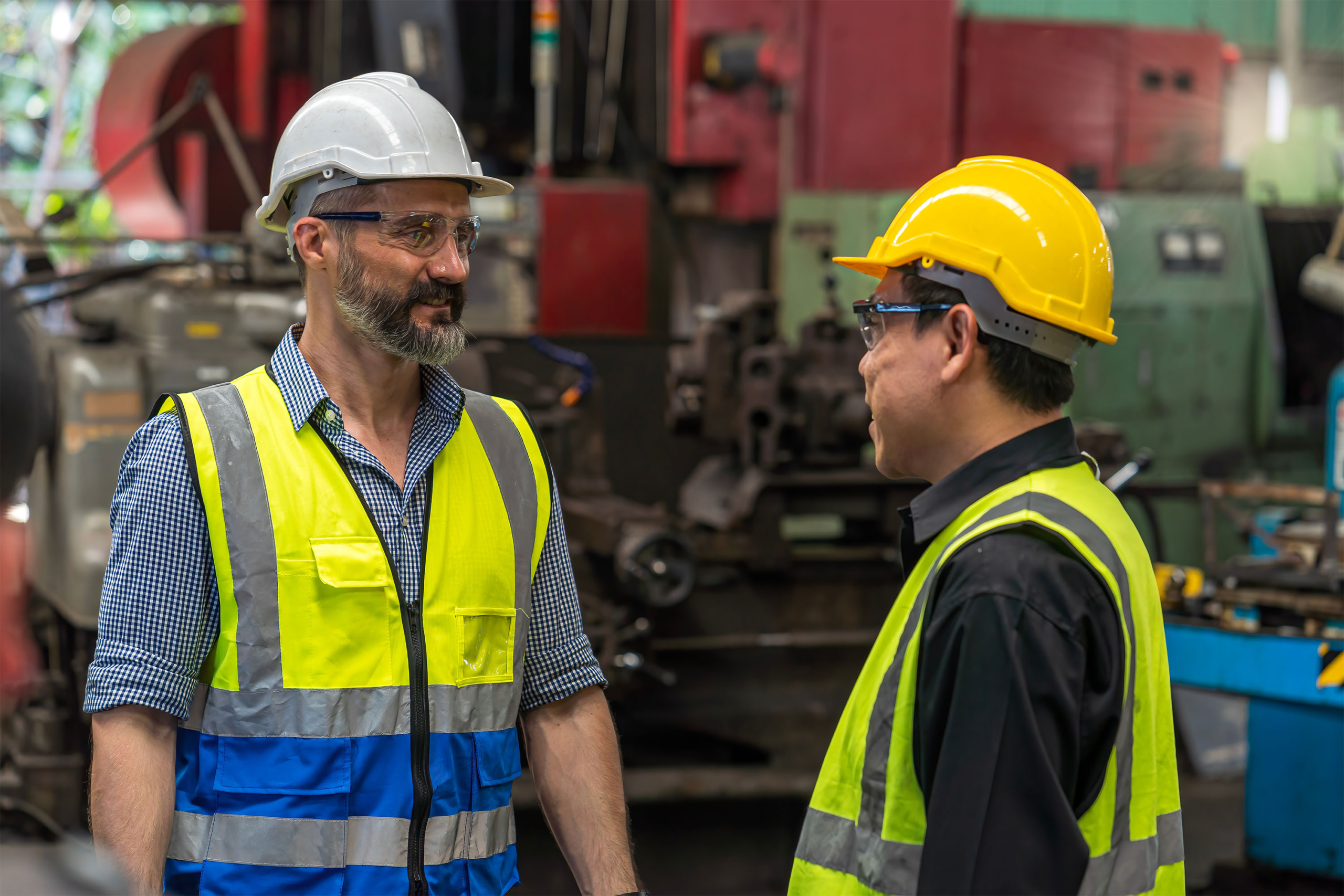 Two people in hard hat, safety glasses, and high-visibility vest talk in a factory