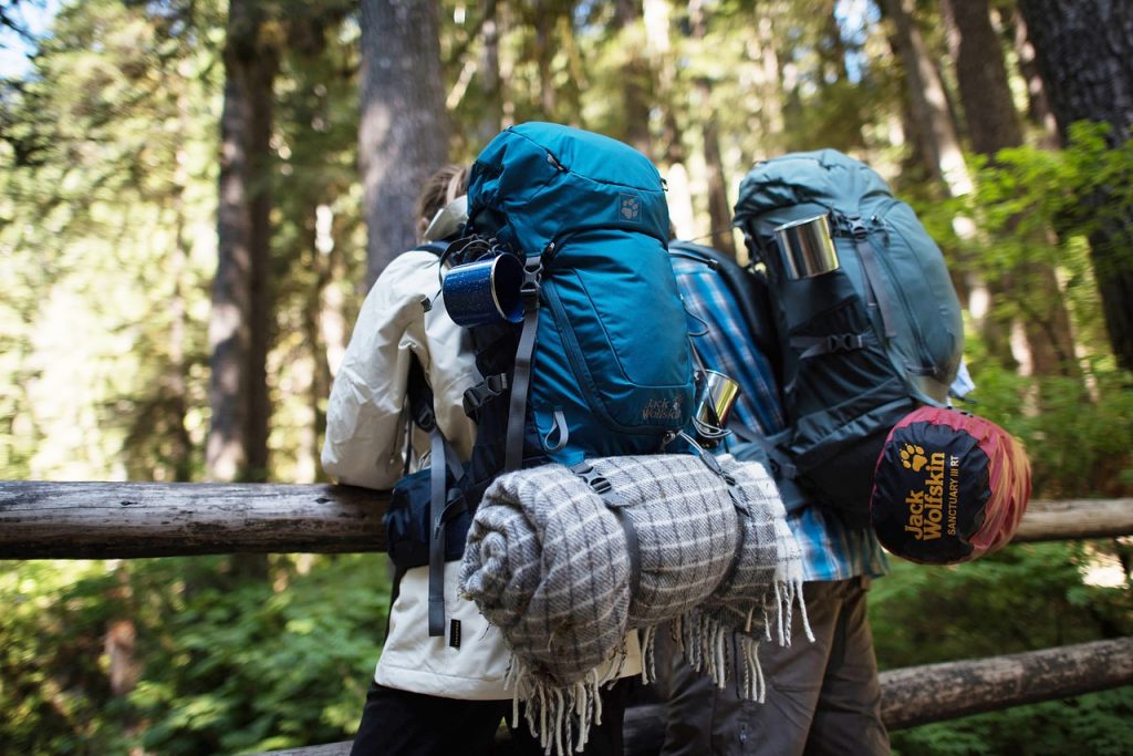 The Best Smart Backpacks You Can Get Right Now Two hikers with Jack Wolfskin backpacks and camping gear standing in a sunny forest, focusing on outdoor equipment.