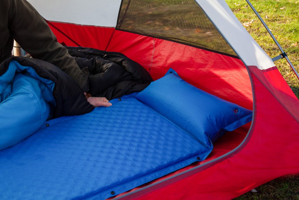 A man gets his tent and sleeping bag ready at a campground by inflating and setting up his blue blow-up mattress pad to put for under his sleeping bag