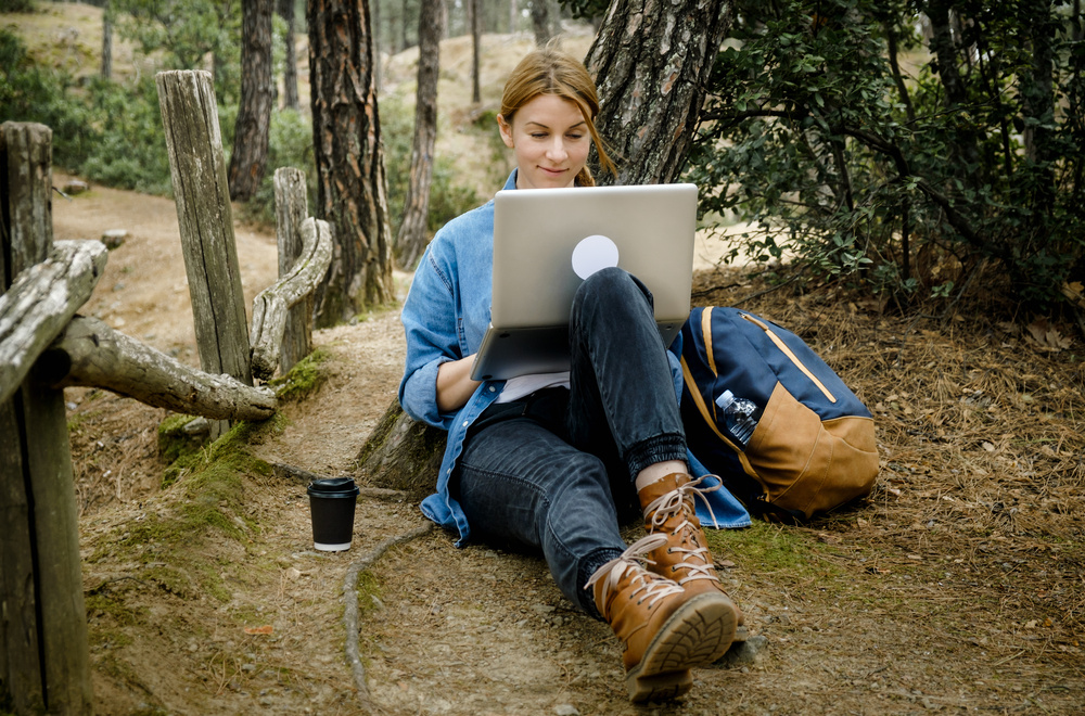 Young woman with backpack sitting on ground at forest and working on laptop . Workation concept. Freelancer work