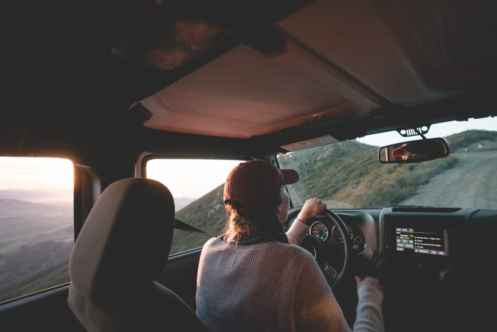 View from the back seat of a 4x4 vehicle showing a driver in a baseball cap and sweater driving on a steep, unpaved mountain road during sunset, with an orange glow visible through the window.