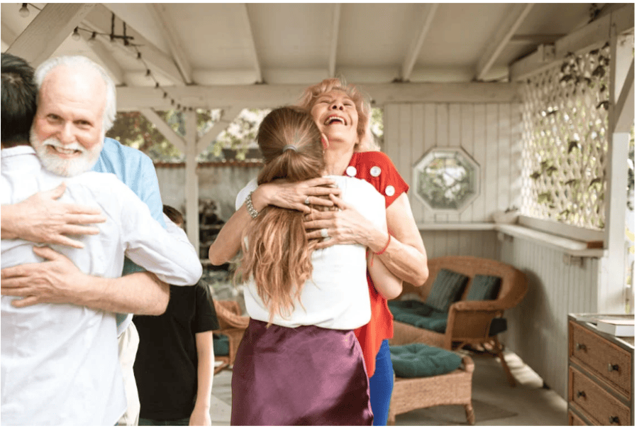 An older woman and an older man hug family members on a sunlit outdoor porch during a happy reunion.