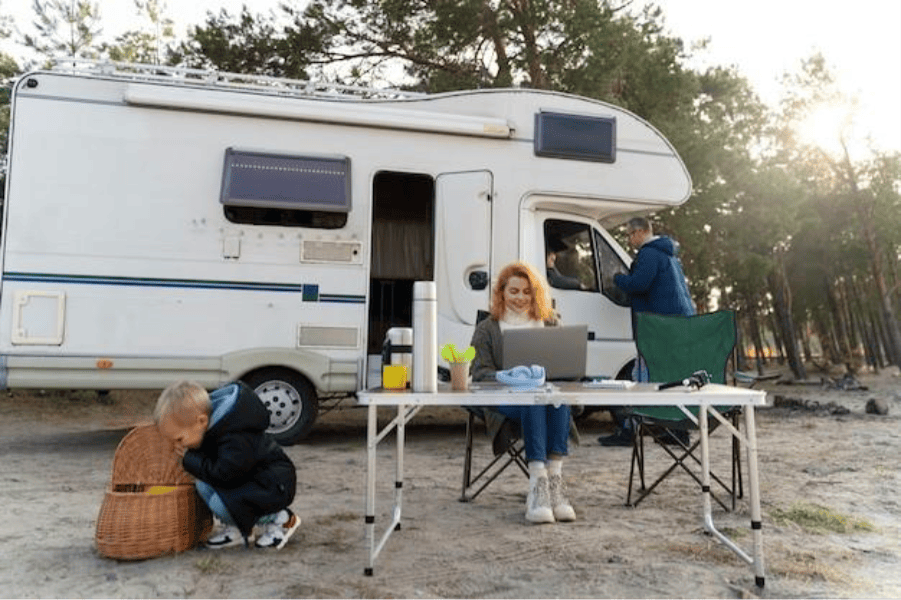 A family camps with their motorhome; a woman works on a laptop at a table while a child plays with a basket nearby.
