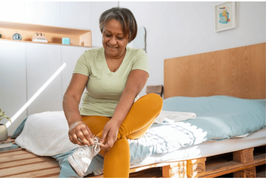 7 Benefits of Traveling Solo When Over 30 An older woman with a smile on her face sits on a bed and ties the laces of her white sneakers.