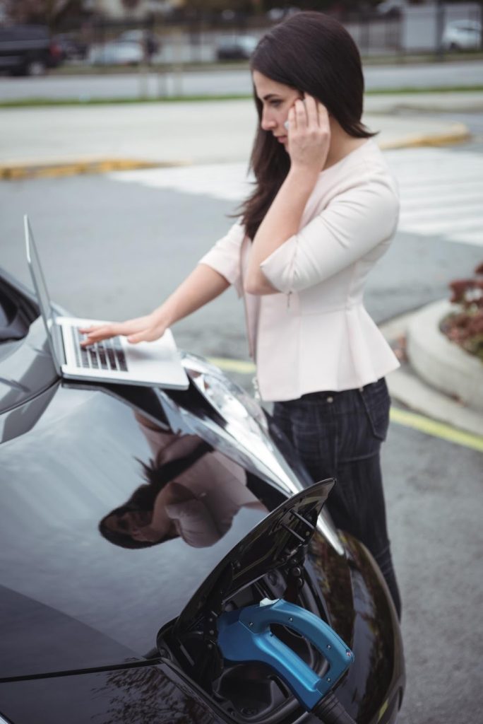 A woman stands next to her black electric car as it charges, talking on her phone while working on a laptop on the car's hood.