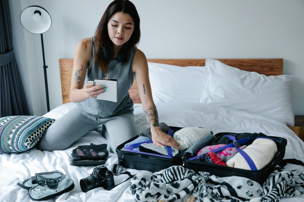 A young woman is kneeling on a bed, consulting a packing list while organizing clothes and a camera into an open suitcase.