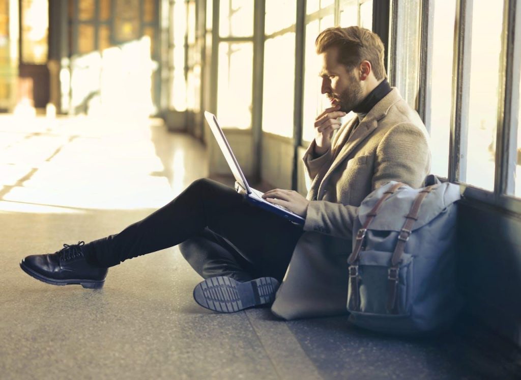 Stylish man sitting on the floor with a backpack, working intently on a laptop in a bright, sunlit hallway or terminal.