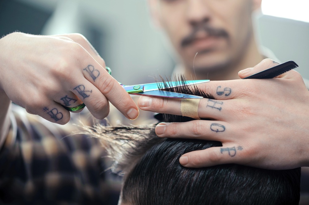 Close-up of a barber with tattooed knuckles (BARB SHOP) using iridescent scissors and a comb to cut a client's hair.