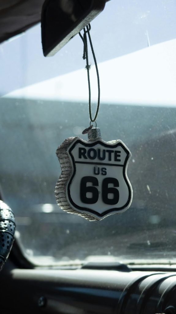 Close-up of a white and black Route US 66 shield ornament hanging from the rearview mirror inside a car, with the bright, blurry view of the road visible through the windshield.