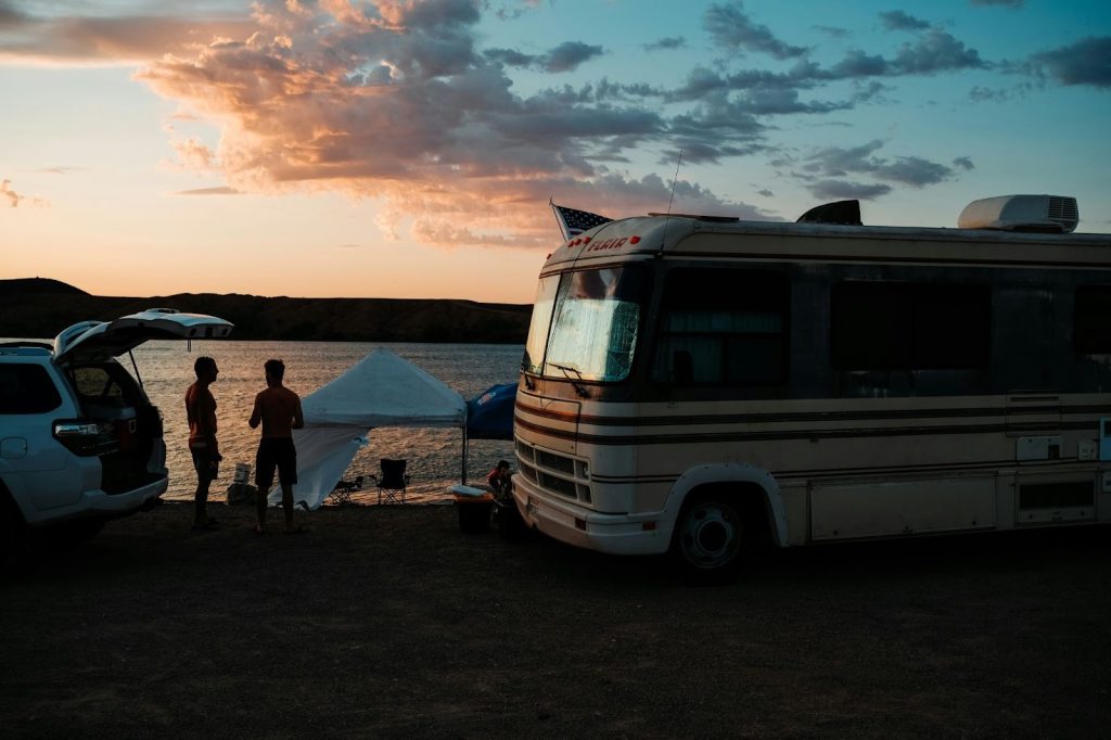 RV and SUV parked by a lake at sunset with two people setting up a tent for camping.