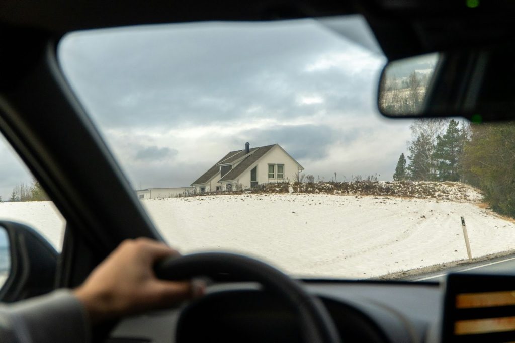 View from inside a car of a white house on a snowy hillside under a cloudy sky.