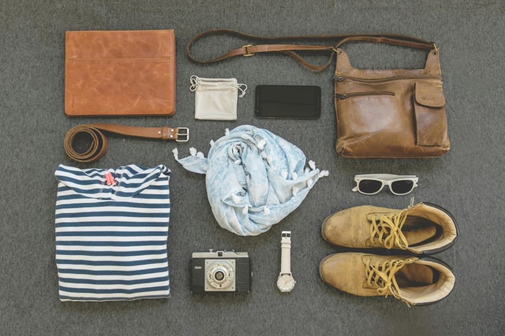 Flat lay of travel essentials including clothes, boots, camera, sunglasses, and leather accessories on a gray surface.