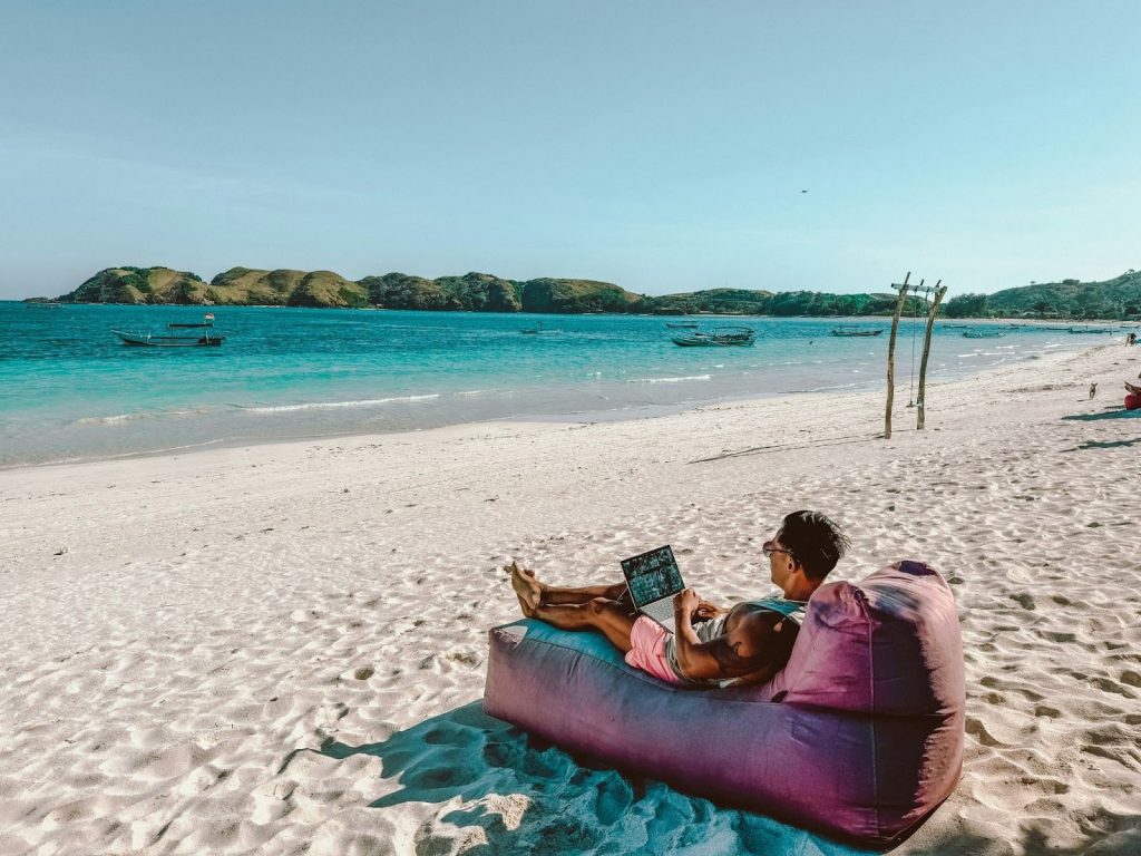 Man working on a laptop while relaxing on an inflatable lounger on a sunny tropical beach with turquoise water.