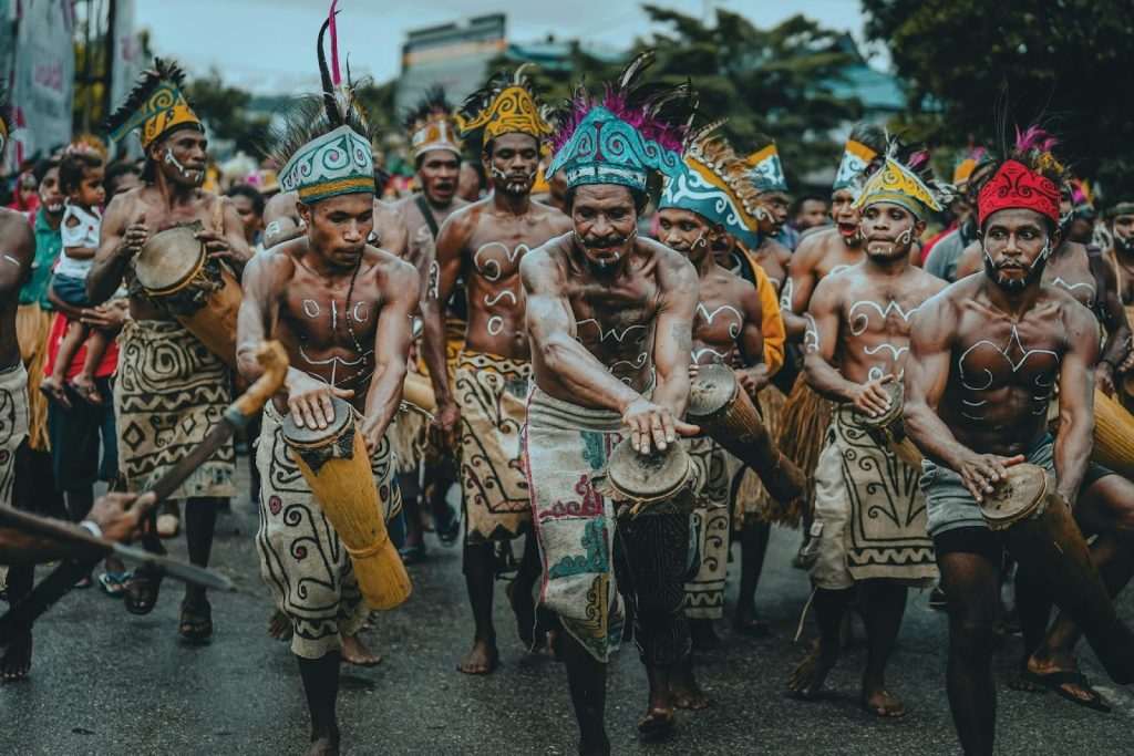 Group of men in colorful feathered headdresses and body paint playing drums during a cultural procession in Indonesia