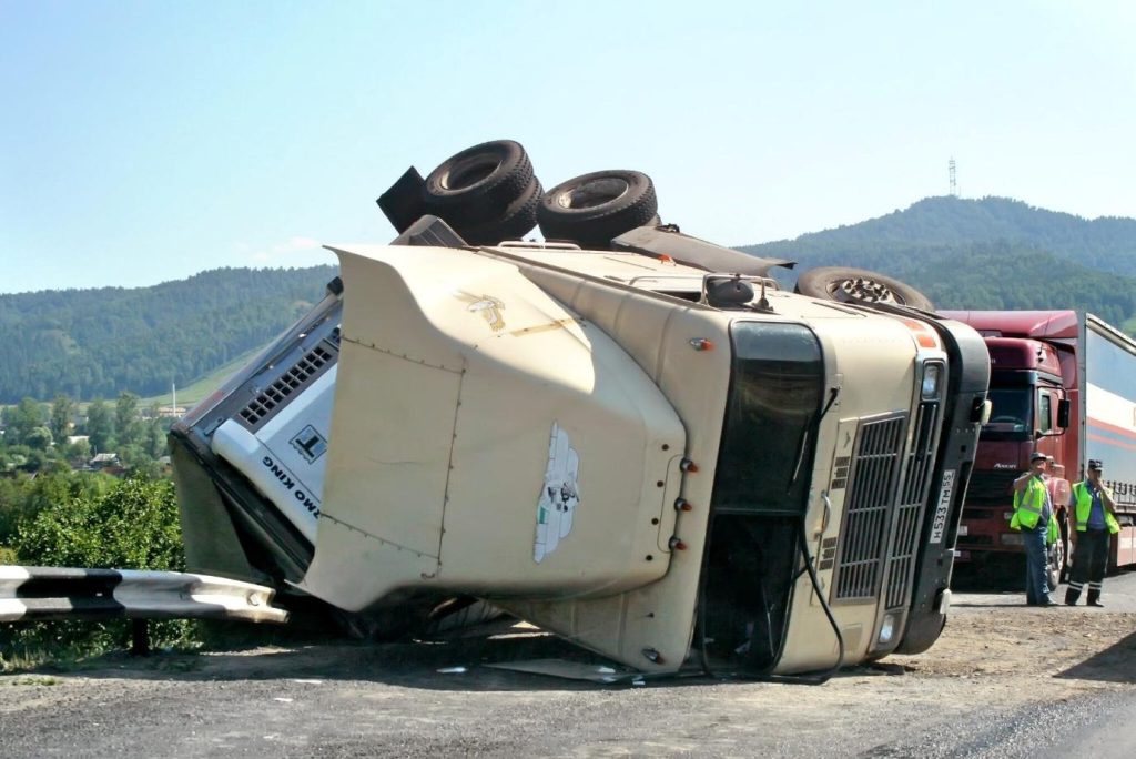 Overturned semi-truck tractor blocking a mountain highway with recovery workers on a sunny day.