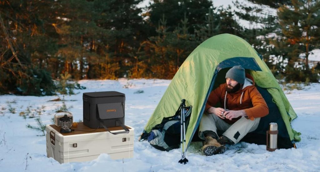 Man camping in a green tent in the snow with a Jackery portable power station and camping gear.