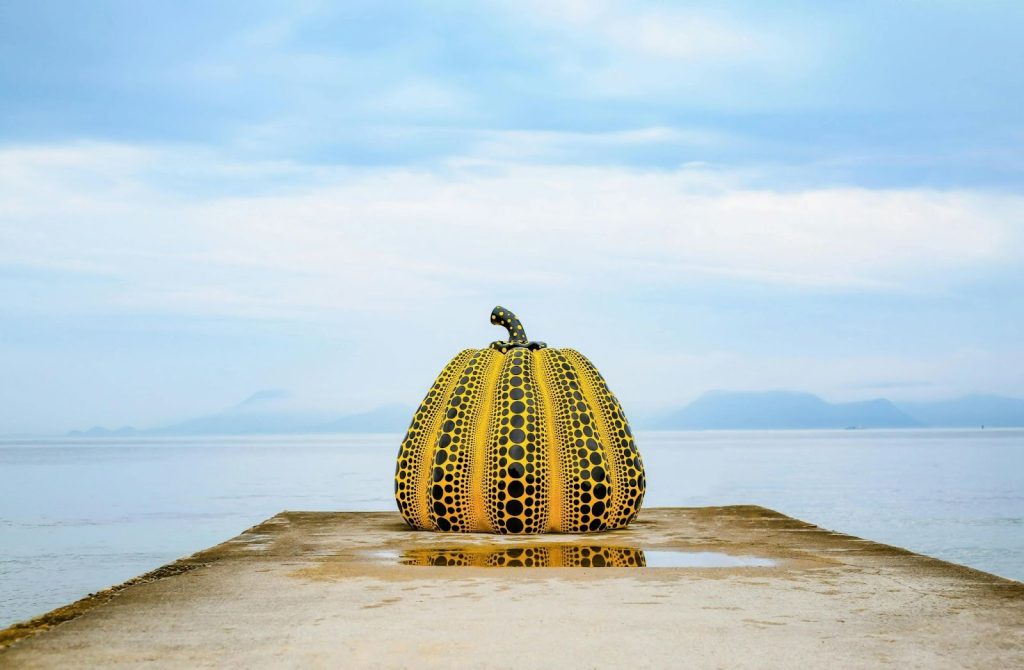 Yayoi Kusama's Yellow Pumpkin sculpture with black polka dots on a pier overlooking the ocean.
