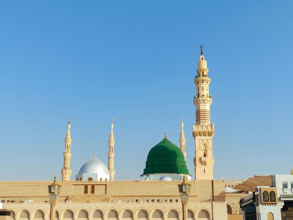 Prophet's Mosque in Medina, Saudi Arabia, showing the Green Dome and an ornate minaret against a blue sky.